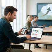 Un homme travaillant sur un ordinateur portable et un smartphone dans un bureau moderne avec des collègues en arrière-plan.