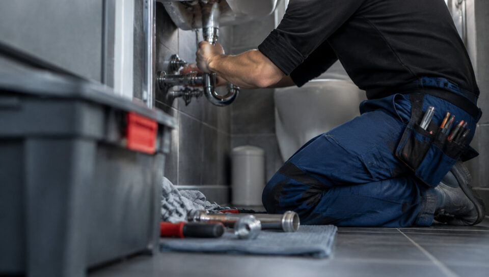 Un plombier professionnel en uniforme agenouillé répare la tuyauterie sous un lavabo dans une salle de bain moderne.